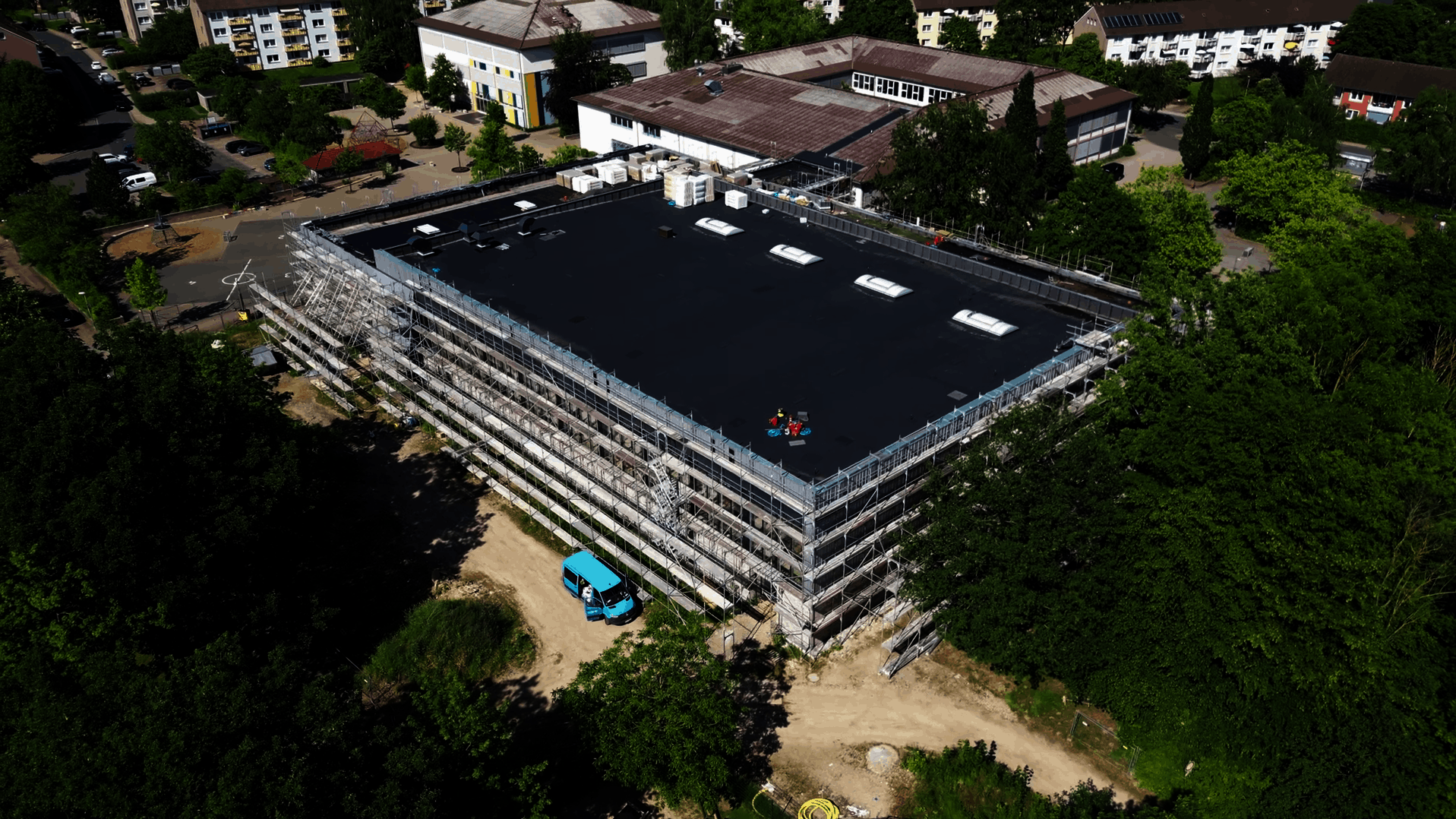 Luftaufnahme einer großen, rechteckigen Sporthalle mit flachem, dunklem Dach und mehreren Oberlichtern. Das Gebäude ist vollständig von einem Baugerüst umgeben. Die Halle ist von Bäumen und weiteren Gebäuden umgeben. Es handelt sich um die Sporthalle Barne in Wunstorf, erbaut von Gundlach Bauunternehmen.