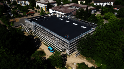 Luftaufnahme einer großen, rechteckigen Sporthalle mit flachem, dunklem Dach und mehreren Oberlichtern. Das Gebäude ist vollständig von einem Baugerüst umgeben. Die Halle ist von Bäumen und weiteren Gebäuden umgeben. Es handelt sich um die Sporthalle Barne in Wunstorf, erbaut von Gundlach Bauunternehmen.