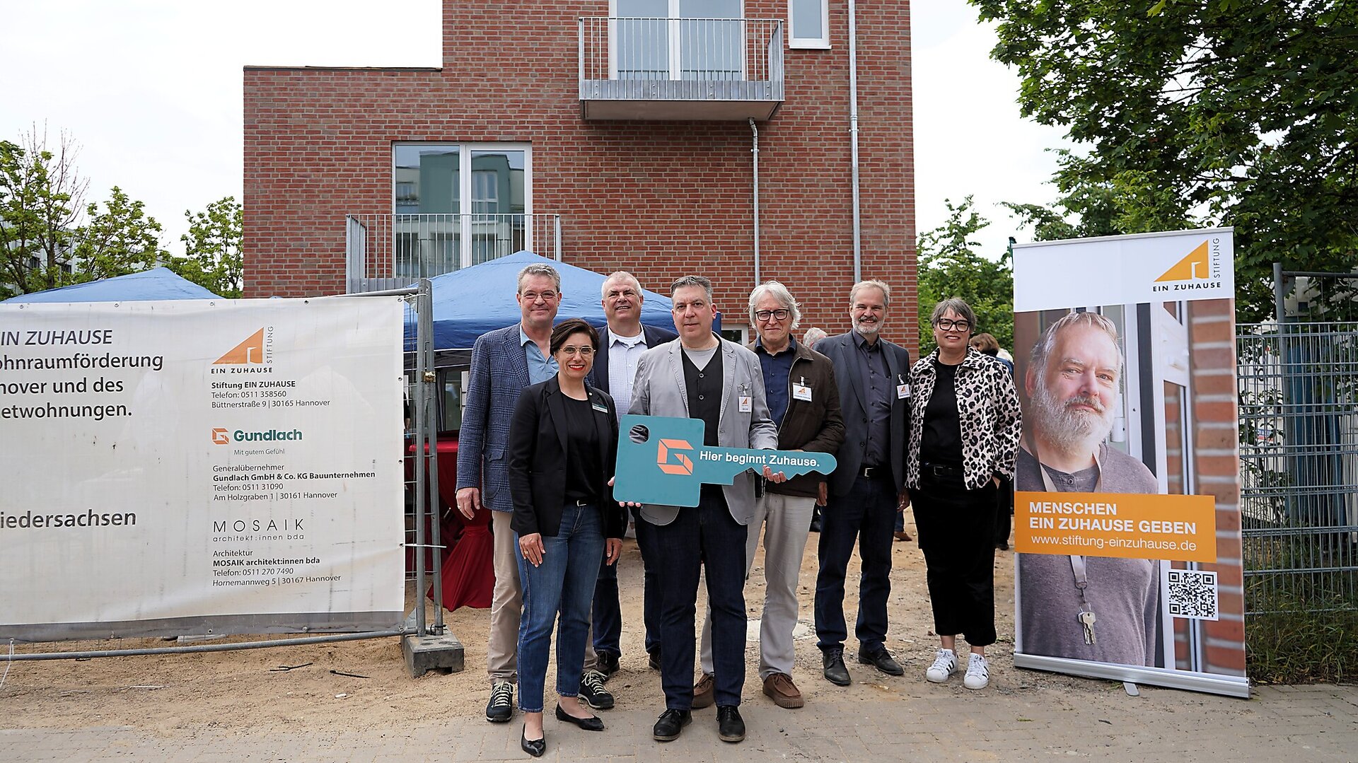 Nadine Otto übergibt für das Gundlach Bauunternehmen den Schlüssel mit dem Text 'Hier beginnt Zuhause'. Das Bauprojekt für Menschen in Wohnungsnot umfasst 7 Wohneinheiten. Schlüsselfertig gebaut.