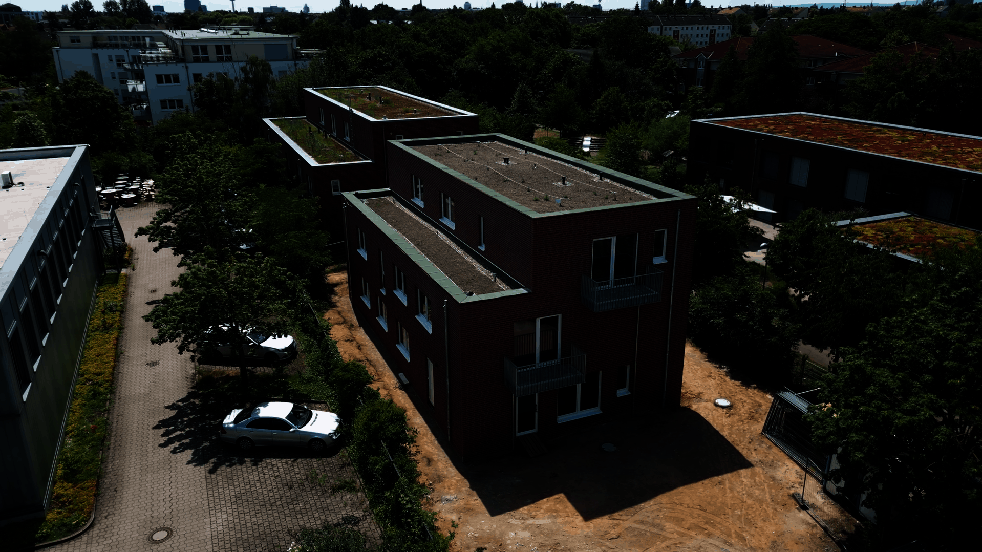 Im Mittelpunkt steht ein modernes, zweistöckiges Backsteingebäude mit Flachdach und mehreren kleinen Balkonen. Das Gebäude ist Teil des Wohnprojekts »Housing First« für Menschen in Not am Karl-Imhoff-Weg. Die Umgebung ist grün, mit Bäumen und weiteren Gebäuden, von denen einige begrünte Dächer haben. Ein weißes Auto steht auf einem Parkplatz neben dem Haus. Die Szene wirkt ruhig und gepflegt.