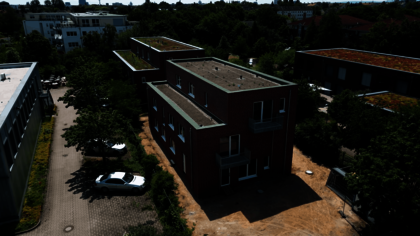 Im Mittelpunkt steht ein modernes, zweistöckiges Backsteingebäude mit Flachdach und mehreren kleinen Balkonen. Das Gebäude ist Teil des Wohnprojekts »Housing First« für Menschen in Not am Karl-Imhoff-Weg. Die Umgebung ist grün, mit Bäumen und weiteren Gebäuden, von denen einige begrünte Dächer haben. Ein weißes Auto steht auf einem Parkplatz neben dem Haus. Die Szene wirkt ruhig und gepflegt.