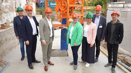 Im Mittelpunkt steht eine Gruppe von Menschen, die auf einer Baustelle zusammenkommt - darunter Nadine Otto und Belit Onay. Sie tragen orange Bauhelme mit einem weißen „G“ des Gundlach Bauunternehmes und stehen um eine frisch gegossene Betonfläche. Im Hintergrund sind eine orange Betonmischmaschine, Baucontainer und Gebäudeteile zu sehen. Die Szene zeigt die feierliche Grundsteinlegung für Micro Apartments für ehemals wohnungslose Menschen an der Büttnerstraße gebaut durch das Bauunternehmen Gundlach.