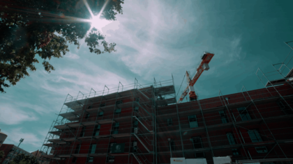 Ein mehrstöckiges Backsteingebäude mit Gerüsten an der Fassade steht im Vordergrund. Im Hintergrund ragt ein Baukran über das Gebäude. Sonnenlicht fällt durch die Baumkrone am linken oberen Bildrand. Die Szene zeigt den Neubau von Micro Apartments für ehemals wohnungslose Menschen in der Büttnerstraße. Das ausführende Bauunternehmen ist das Gundlach Bauunternehmen.